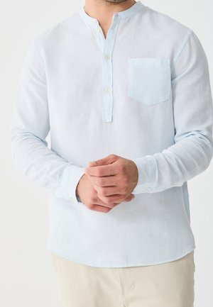 Man draagt een lichtblauw Henley-shirt met lange mouwen, borstzakje, knoopkraag en beige broek, handen gevouwen voor zich.