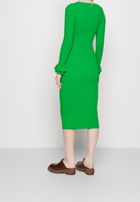 Grünes, geripptes Kleid mit langen Ärmeln, Rüschenbündchen und einem Reißverschluss hinten. Getragen mit braunen Clogs, knielang und figurbetont.