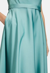 Ein türkisfarbensatin Kleid mit tailliertem Oberteil, Taillenschnürung und vollem Rock mit Taschen, das eine glatte, leicht reflektierende Textur zeigt.