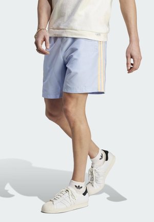 Mężczyzna w jasnoniebieskich szortach z żółtymi paskami po bokach, beżowej koszuli, białych butach Adidas i białych skarpetkach, stojący z jedną nogą skrzyżowaną.