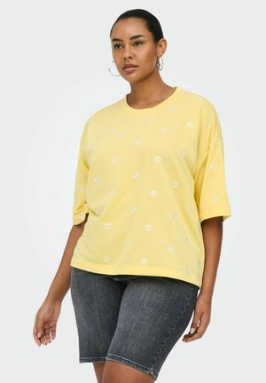 Kvinde iført gul oversized T-shirt med hvide blomsterprint og grå denim knælange shorts, stående mod en hvid baggrund.