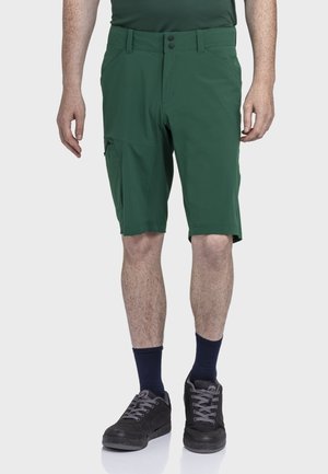 Mann trägt dunkelgrüne kniehohe Shorts, dunkle Socken und schwarze Turnschuhe, steht vor einem schlichten hellgrauen Hintergrund.