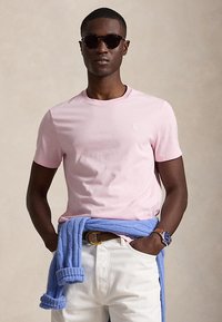 Man wearing black sunglasses, pink t-shirt, white pants, blue sweater tied around waist, and wristwatch with brown leather strap.