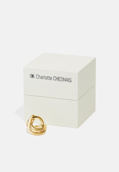 Valge Charlotte Chesnais ehtekast musta tekstiga ja skulpturaalne kuldsõrmus selle ees valgel pinnal.