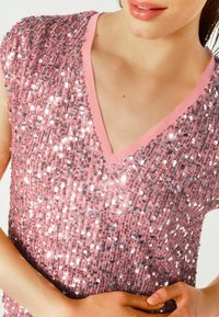 Růžový top se známkami s výstřihem do V, pokrytý lesklými stříbrnými flitry na měkkém povrchu, s krátkými rukávy a růžovým lemem kolem výstřihu.