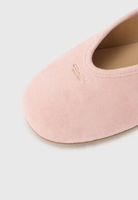 Ροζ σουέτ παπούτσι slip-on με λεία υφή, στρογγυλεμένη μύτη και χρυσό λογότυπο "COACH" στο πλάι. Μινιμαλιστικός σχεδιασμός,  χωρίς ορατά μεταλλικά μέρη.