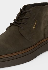 Bottine en daim marron foncé avec lacets assortis, semelle marron avec coutures et logo doré "GANT" sur le côté.