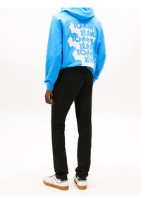 Modrá mikina s kapucí a velkým bílým grafickým textem "TOMMY JEANS" na zadní straně, spárovaná s černými kalhotami a bílými teniskami s černými akcenty.