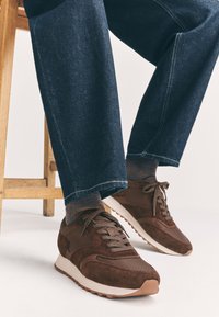 Chaussures de sport marron en mélange de suède et de matériaux synthétiques, avec une tige à motif, des lacets ronds et une semelle en caoutchouc blanche.