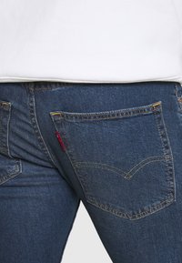 Blå denimjeans bakficka med gula stygn och en liten röd tygetikett, bärs med en vit skjorta.