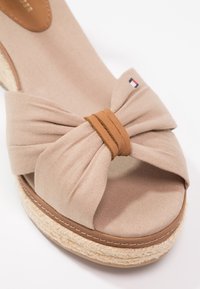Sandale compensée beige à bout ouvert avec un nœud en tissu et un détail en cuir enroulé, ornée d'un petit emblème de drapeau rouge, blanc et bleu sur le nœud.