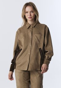 Jonge vrouw draagt een losse bruine suède overhemd met knopen en donkere zijpanelen, met bijpassende broek, staande tegen een effen lichte achtergrond.