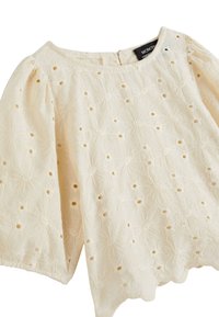 Blouse crème avec un design floral en œillets, ourlet en forme de vagues et manches bouffantes. Présente une fermeture à boutons au dos et un tissu doux et texturé.