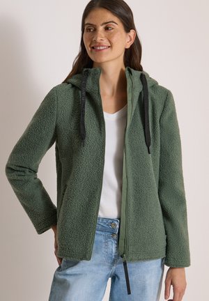 Mujer sonriendo, lleva una chaqueta de forro polar verde con capucha y cremallera, camisa blanca y vaqueros azul claro, de pie con una mano en la cadera.