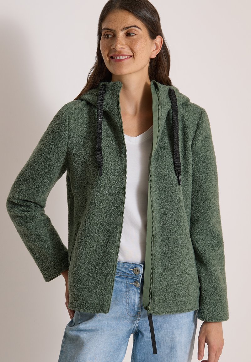 Femme souriante portant une veste polaire verte à capuche zippée, une chemise blanche et un jean bleu clair, debout avec une main sur la hanche.