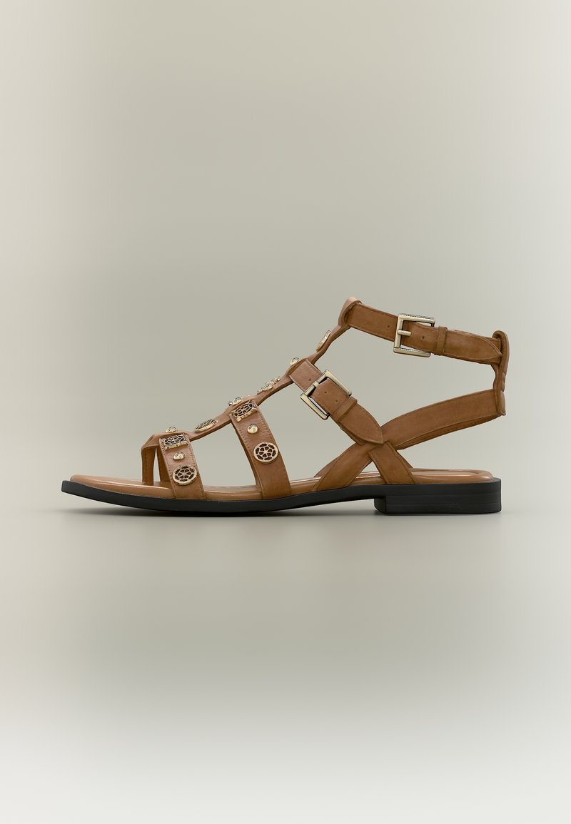 Καφέ δερμάτινο επίπεδο σανδάλι γλαδιόλα με λουριά στον αστράγαλο, μεταλλικές αγκράφες και διακοσμητικά στρογγυλά καρφιά σε ουδέτερο φόντο.