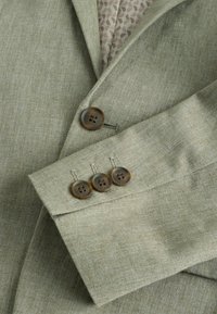 Veste blazer vert clair texturée avec quatre boutons marron sur la manche et un bouton fermé à l'avant sur une chemise beige à motifs.