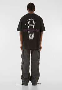 Sort bomulds t-shirt med et stort grafisk print af et hundehoved med åben mund på ryggen. Løs pasform med korte ærmer.