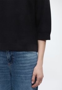 Δεξί χέρι και κορμός με μαύρο ριμπ τριών τετάρτων μανίκι και μεσαίου ξεβγαλμένου μπλε τζιν παντελόνι, σε λευκό φόντο.