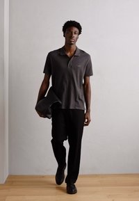 Grå polo skjorte med et subtilt design, parret med sorte bukser. Bærer en sort lædertaske, står på et trægulv mod en neutral baggrund.
