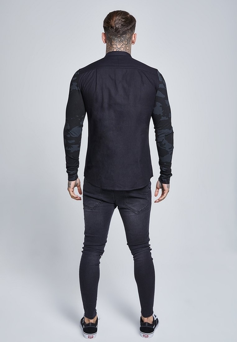 Homme avec un grand tatouage au cou portant une chemise noire à manches longues avec des manches camouflage, jean noir skinny et baskets noires, debout de dos.