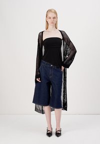 Cardigan preto transparente de malha sobre um top preto sem alças, combinado com culottes de ganga azul soltas e sapatos de salto agudo pretos.