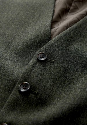 Sötétzöld gyapjú szövet sima textúrával, két fekete gombbal és barna mintás béléssel. Közeli nézet a ruházati részletekről.