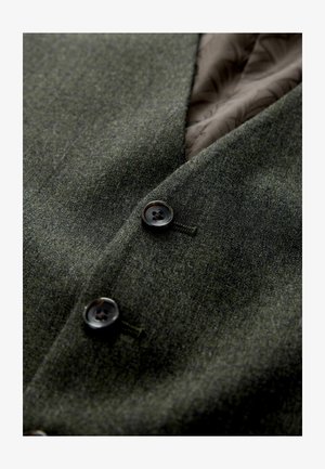 Sötétzöld gyapjú szövet sima textúrával, két fekete gombbal és barna mintás béléssel. Közeli nézet a ruházati részletekről.