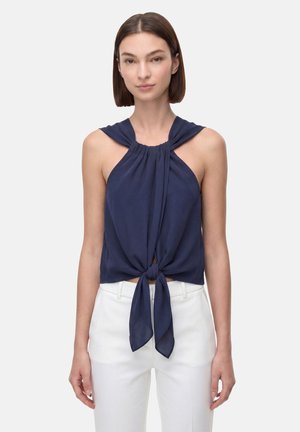 Donna che indossa un top smanicato blu navy halter annodato in vita e pantaloni bianchi a vita alta, in piedi davanti a uno sfondo semplice.