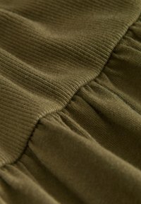 Nærbillede af ribbet olivengrønt stof med en tekstureret overflade og detaljer med samlet stof, der skaber et blødt, lagdelt udseende.