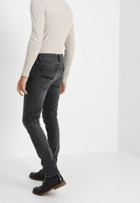 Úzké černé džíny s jemnou texturou, obsahující pět kapes a zakřivený šev na zadní kapse, doplněné černými botami.