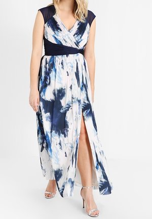 Woman in sleeveless blue and white floral maxi dress with wrap neckline and thigh-high slit, wearing silver high-heeled sandals.
