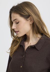 Jeune femme aux cheveux châtain clair, portant une chemise marron foncé boutonnée, regardant vers le bas sur un fond blanc uni.