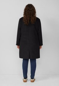 Zwarte jas met een rechte snit, lange mouwen en een split aan de achterkant. Draagend over blauwe jeans en sneakers, gezien vanuit de achterkant.