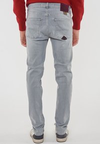 Mann in hellblauen Jeans, rotem Langarmshirt und weißen Turnschuhen, stehend mit dem Rücken zu einer neutralen Hintergrundwand.
