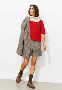 Pull rouge à manches courtes superposé à un col beige, associé à un short à carreaux, un blazer gris à carreaux, des chaussures marron à motif léopard et des chaussettes texturées.