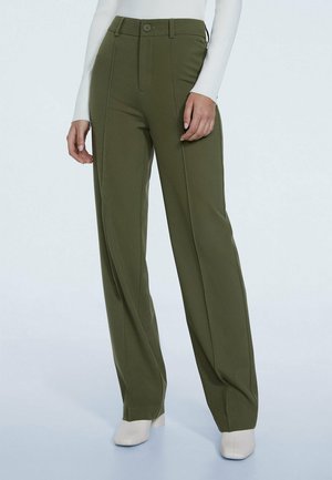 Hohe Taille, olivgrüne Hose mit weitem Bein, vordere Nähte, Knopf- und Reißverschluss, kombiniert mit weißen Knöchelstiefeln und einem weißen Langarmshirt.