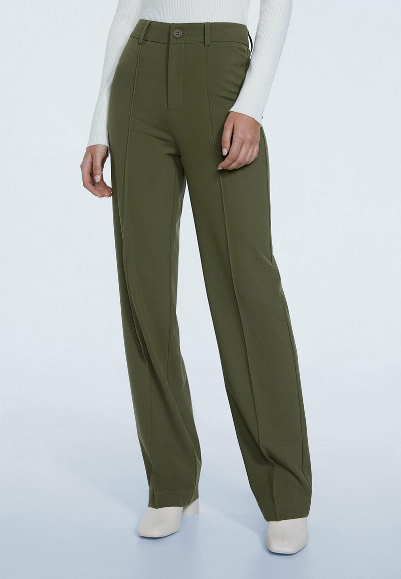 Pantalon taille haute vert olive à jambes larges avec coutures devant, fermeture bouton et zip, porté avec des bottines blanches et un haut blanc à manches longues.