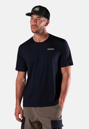 Uomo che indossa una t-shirt nera Rossignol e un cappello a rete nero, in piedi con una mano nella tasca dei pantaloni cargo color kaki, su uno sfondo chiaro e uniforme.