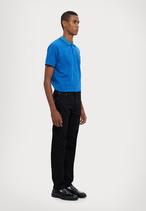 Blå polo med et subtilt mønster, grøn krave, parret med sorte bukser og sorte sko. Snittet er slankt og moderne.