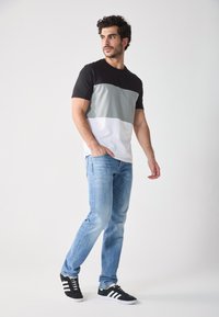 Tricou colorat în blocuri de negru, gri și alb, asortat cu blugi albaștri deschiși și adidași negri cu accente albe. Material neted, croi lejer.