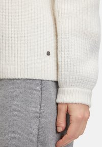 Gestrickter cremfarbener Pullover mit einem strukturierten Muster, ausgestattet mit gerippten Bündchen und einem kleinen silbernen Druckknopf-Detail. Kombiniert mit einer grauen, strukturierten Hose.