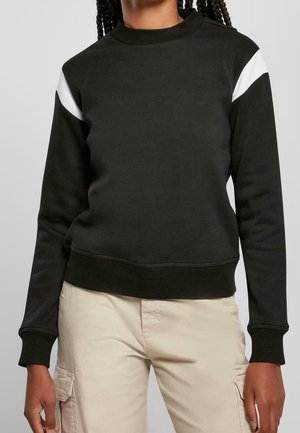 Person iført en sort langærmet sweatshirt med hvide diagonale striber på skuldrene og beige cargobukser, stående mod en grå baggrund.