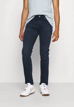 Persona che indossa jeans slim fit blu scuro, camicia grigio chiaro infilata e sneaker bianche con swoosh beige, in piedi su pavimento grigio chiaro.