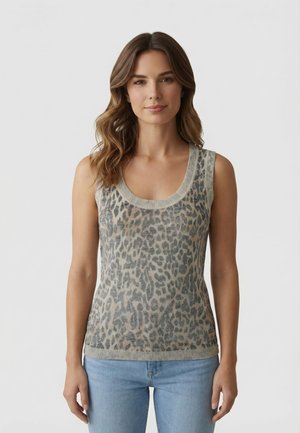 Donna con capelli castano ondulati indossa un top smanicato con scollo a barca e stampa leopardata, e jeans azzurri, in piedi contro uno sfondo chiaro e semplice.