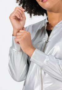 Stříbrná bomber bunda se lesklým metalickým povrchem, zapínání na zip a zipovým táhlem ve tvaru hvězdy. Rukávy jsou na manžetách stažené.