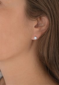 Strieborné náušnice vo forme srdca so malým čírym drahokamom, umiestnené proti hladkému, prirodzenému tónu pleti. Jednoduchý, elegantný dizajn.