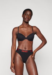 Soutien-gorge noir à fines bretelles et texture lisse, associé à un slip de bikini noir avec une taille "HUGO" rouge.