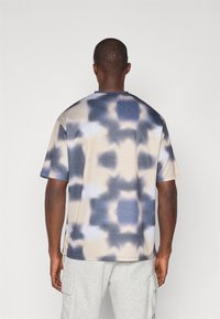 Kortærmet, lyse beige t-shirt med et blåt og gråt tie-dye mønster, lavet af et struktureret stof med en rund halsudskæring.