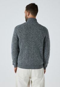 Grau gestrickter Pullover mit hohem Kragen, geripptem Muster und langen Ärmeln. Sichtbare Nähedetails und eng anliegende Bündchen am Saum.